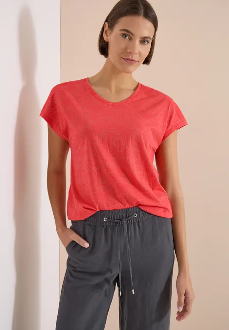 Shirt met ton-sur-ton burn-outpatroon coral Frau in pinkem T-Shirt und grauen Jogginghosen vor hellem Hintergrund.