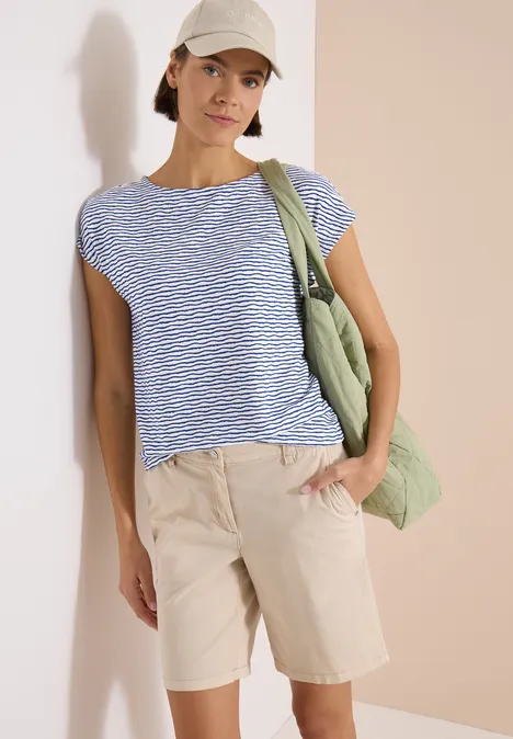 Femme en t-shirt bleu à rayures blanches, bermuda beige et sac vert devant un fond neutre.