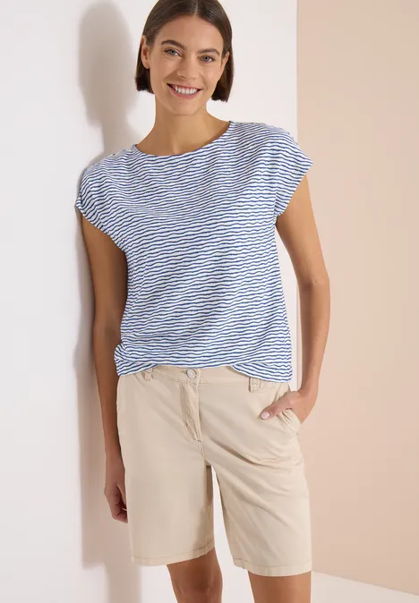 Femme vêtue d’un t-shirt rayé bleu et blanc et d’un short beige, main dans la poche.