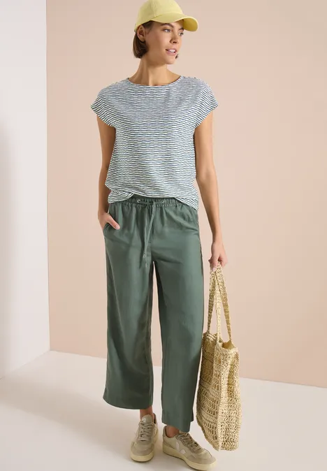 Femme portant un T-shirt rayé, un pantalon large vert, un sac en paille et une casquette.