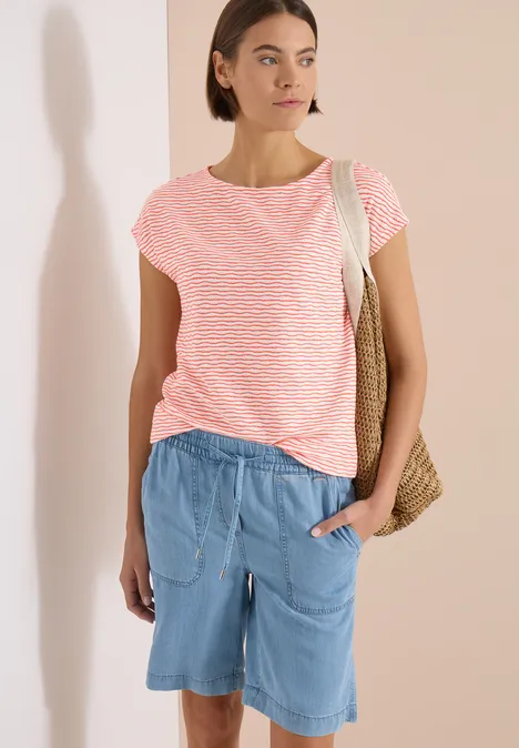 Femme en t-shirt rose à rayures et short bleu, debout devant un fond clair, sac en osier à l'épaule.