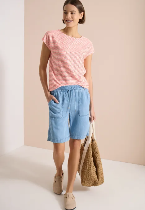 Femme en t-shirt rose, short en jean clair, baskets beiges et sac en osier sur fond neutre.