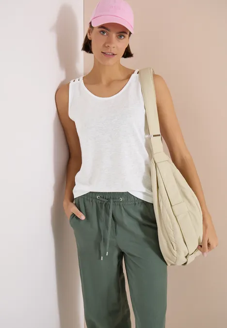 Person lehnt an einer Wand, trägt weißes Tanktop, grüne Hose und große beige Umhängetasche.