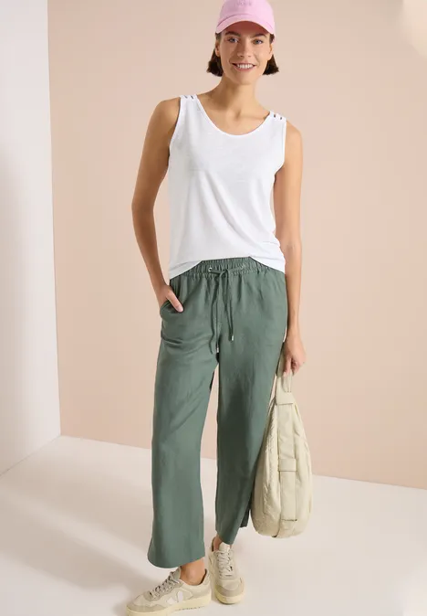 Frau trägt weißes Tanktop, locker sitzende olivgrüne Hose, weiße Sneaker und hält eine helle Stofftasche.
