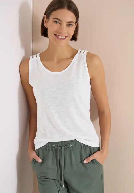Frau trägt weißes Tanktop und grüne Stoffhose, steht mit den Händen in den Taschen vor einer Wand.