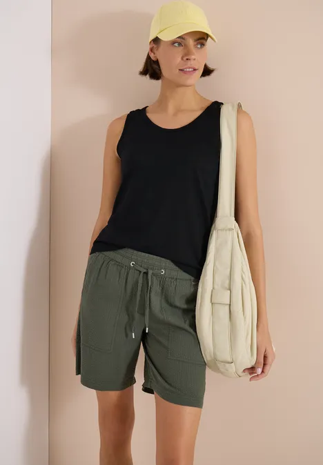 Top avec col en V arrondi et détail tressé Black Femme en débardeur noir, short vert olive et sac beige devant un fond rose.
