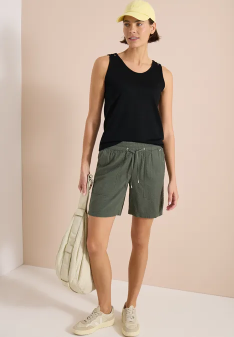 Top avec col en V arrondi et détail tressé Black Personne en débardeur noir, bermuda kaki, baskets beiges et grand sac vert clair devant un fond clair.