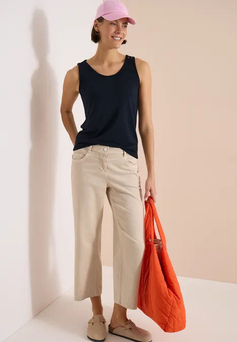 Top avec col en V arrondi et détail tressé universal blue Femme en haut noir et pantalon beige, tenant un sac rouge sur fond clair.