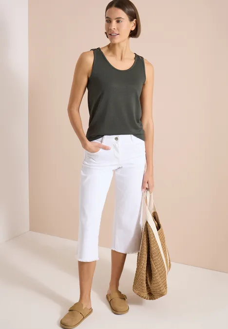 Top avec col en V arrondi et détail tressé khaki Femme en débardeur vert foncé, pantacourts blancs, chaussures beiges et sac en paille sur fond neutre.