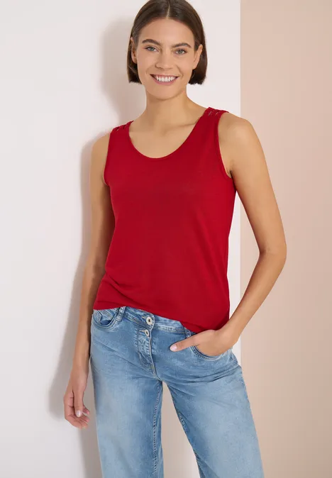 Top avec col en V arrondi et détail tressé tamarillo red Femme en débardeur rouge et jean bleu appuyée contre un mur clair.