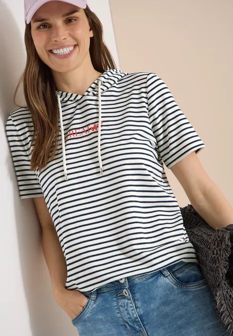 Kurzarm Shirt mit Kapuze und Glitzer vanilla white Frau lehnt entspannt an Wand, trägt gestreiftes T-Shirt mit Kapuze und Jeans, eine Hand in Hosentasche.
