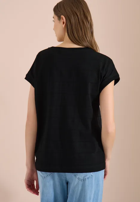 T-shirt structuré avec col bateau Black Frau steht mit dem Rücken zur Kamera, trägt schwarzes T-Shirt und helle, lockere Jeans vor beigem Hintergrund.