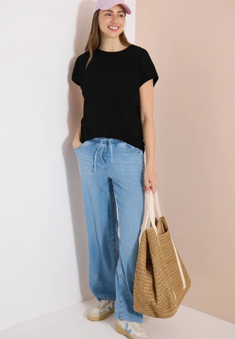 T-shirt structuré avec col bateau Black Frau im schwarzen T-Shirt, weiten hellblauen Hosen und weißen Sneakern, mit geflochtener Strohtasche.