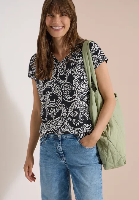 Shirt met V-hals en print Black Vrouw in patroon t-shirt, blauwe jeans en groene gewatteerde tas over de schouder, hand in de zak.