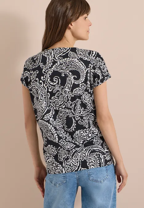 Shirt met V-hals en print Black Vrouw van achteren in blauwe spijkerbroek en zwart-wit paisley t-shirt tegen beige achtergrond.