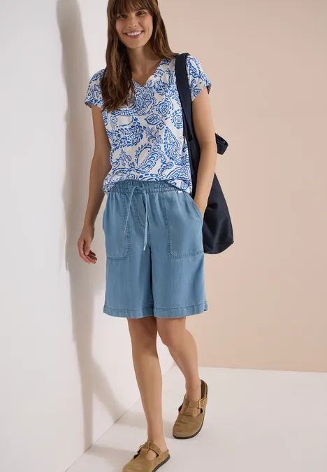 Shirt met V-hals en print vanilla white Vrouw met blauw motief t-shirt, lichtblauwe spijkershorts, kaki sandalen en zwarte tas tegen beige muur