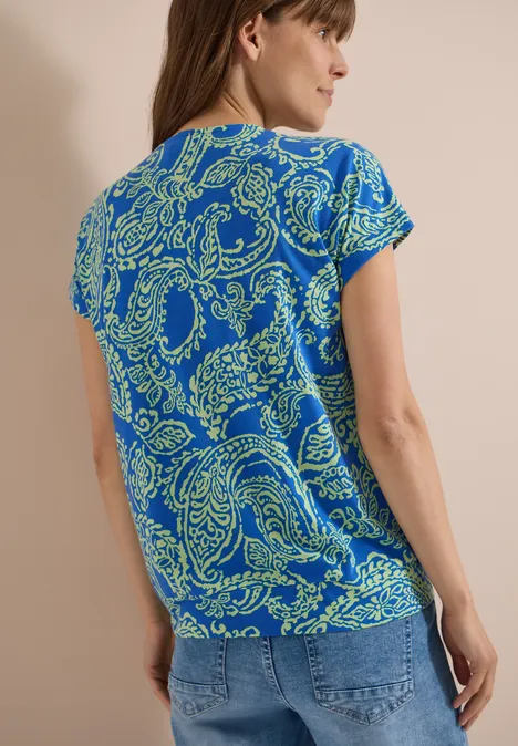 Shirt met V-hals en print surf blue Vrouw van achteren in een blauw t-shirt met groene paisleymotieven en een spijkerbroek.