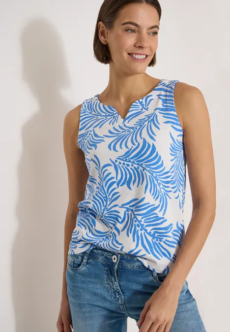 Top met split in de hals en tweekleurig patroon vanilla white Vrouw met mouwloos topje met blauw bladmotief en jeans tegen witte achtergrond.