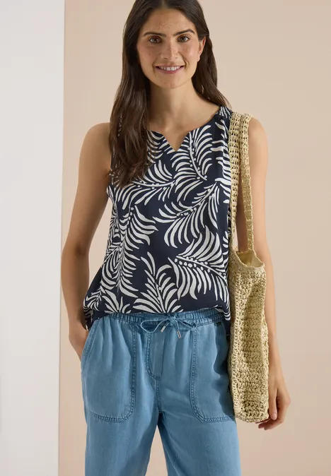 Femme en haut bleu foncé à motif de feuilles, pantalon bleu clair et sac de plage tressé