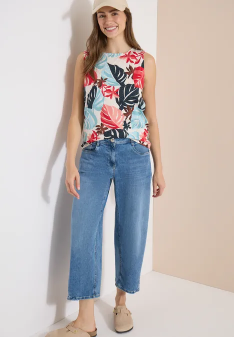 Frau in floralem ärmellosem Top, weiten blauen Culotte-Jeans und beigefarbenen Slippern vor hellem Hintergrund