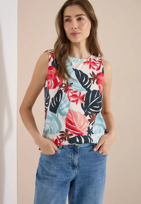 Frau in Jeans und ärmelloser Bluse mit buntem Blatt-Print steht vor neutralem Hintergrund.