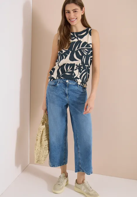 Frau in ärmellosem Top mit Blattmuster, weiten cropped Jeans, hellen Sneakern und gelber Strohtasche.