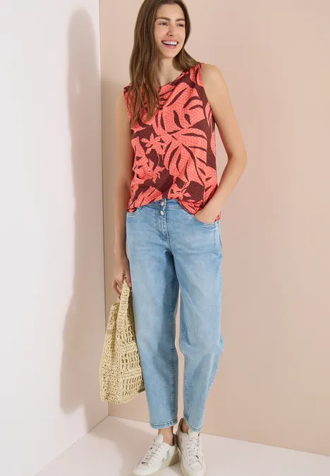 Frau in rotem Top mit Blattmuster, hellblauen Jeans, weißen Sneakers und geflochtener Tasche vor hellem Hintergrund.
