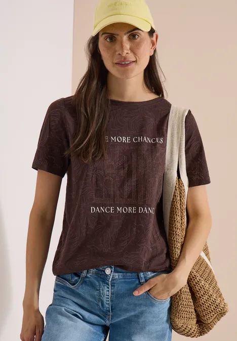 Junge Frau trägt braunes T-Shirt mit Aufdruck, Jeans und Basttasche vor beigem Hintergrund.