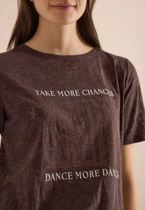 Portrait einer Frau im braunen T-Shirt mit Mustern und den Slogans ‚TAKE MORE CHANCES‘ und ‚DANCE MORE DAYS‘.