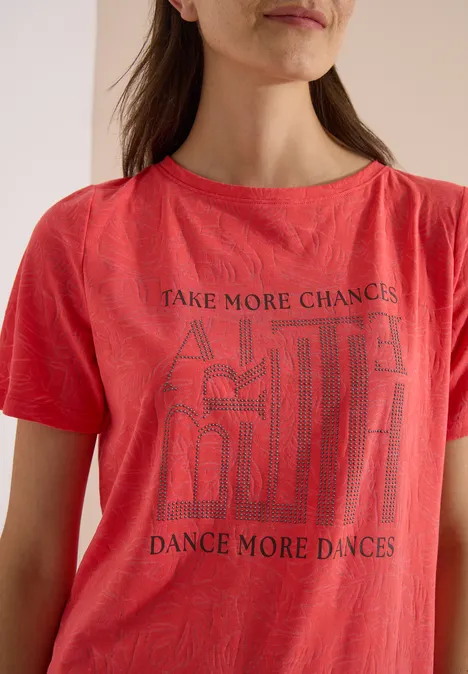 T-Shirt in Ausbrenner-Optik coral Frau in rotem T-Shirt mit Aufdruck 'Take more chances, dance more dances' und abstrakten Linien.