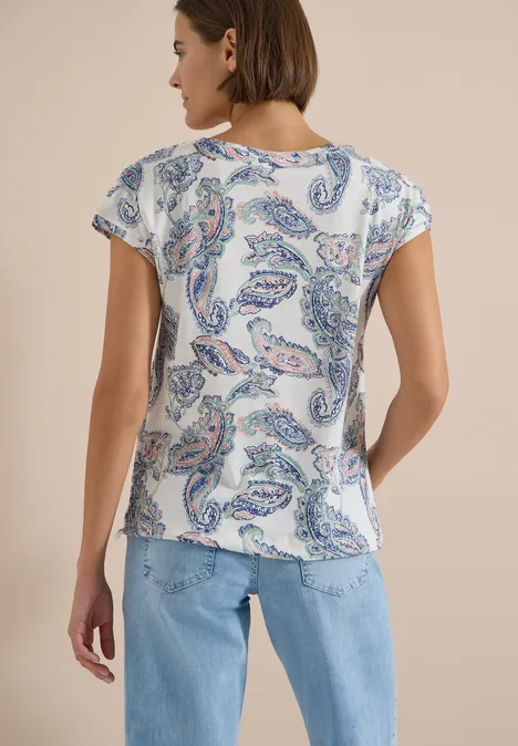 Eine Person von hinten, trägt eine weiße Bluse mit Paisley-Print und blaue Jeans vor beigem Hintergrund.