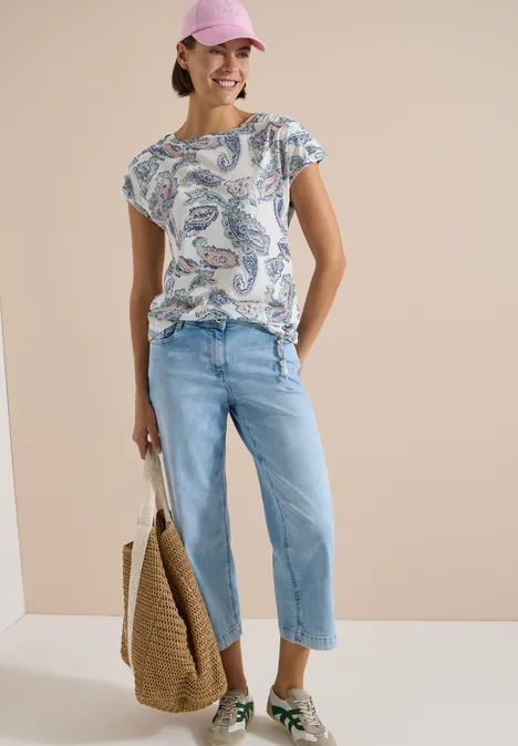 Frau mit gemustertem T-Shirt, hellblauen Cropped-Jeans und Strohtasche vor neutralem Hintergrund.