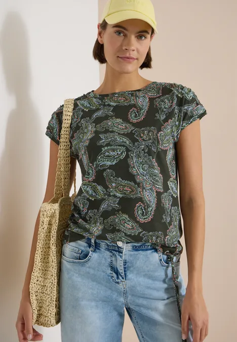 Frau in gemustertem, dunkelgrünem T-Shirt, hellen Jeans und Strohtasche.