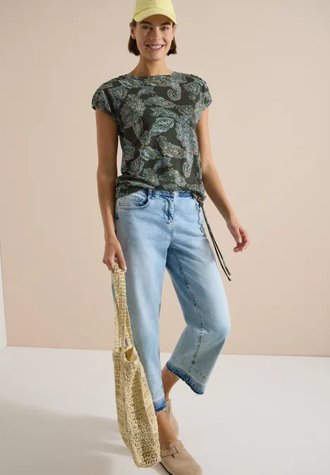 Frau trägt gemustertes Shirt, helle Jeans und hält einen geflochtenen Stoffbeutel vor neutralem Hintergrund.