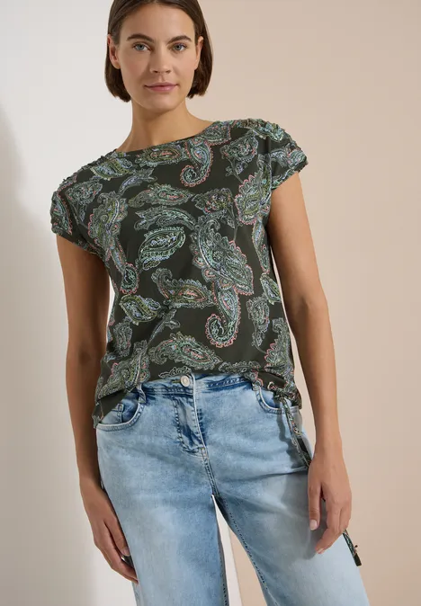 Frau trägt grünes T-Shirt mit Paisley-Muster und lockere blaue Jeans.