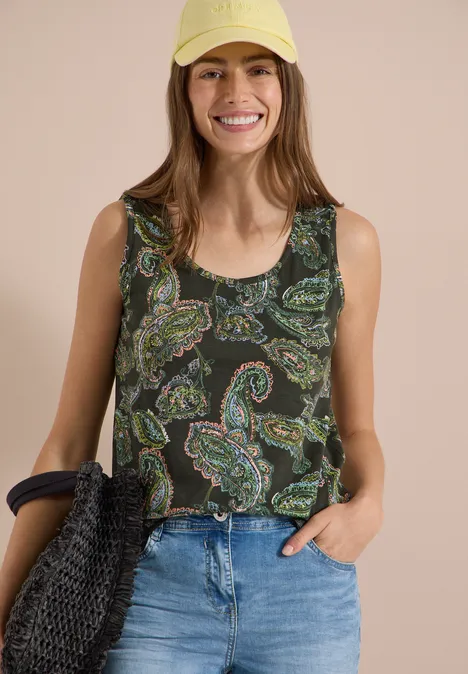 Frau in ärmellosem Paisley-Top und blauen Jeans vor neutralem Hintergrund, hält eine Flechtasche.