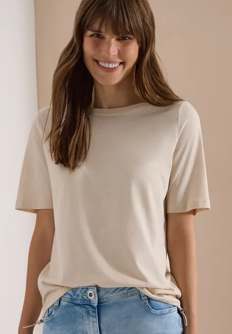 Een persoon draagt een effen beige T-shirt en blauwe spijkerbroek voor een lichte achtergrond.