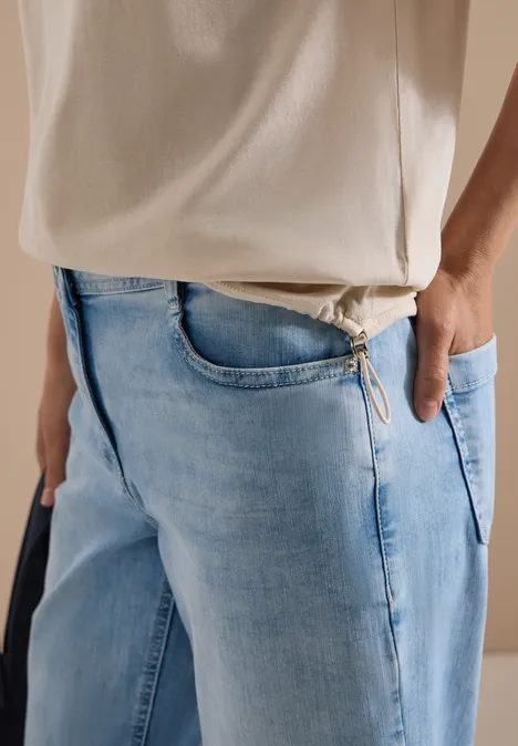Zijaanzicht van een persoon in lichtblauwe jeans en wit T-shirt, hand in zak, zichtbare schouderband.