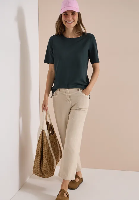 Vrouw voor een beige muur, draagt een donkergroen T-shirt, licht fluwelen broek, rotan tas en bruine sandalen.
