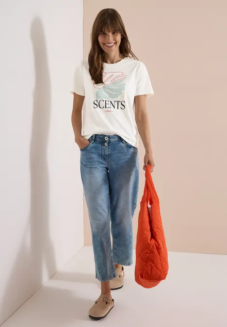 Een vrouw draagt een wit t-shirt met tekst, een wijde spijkerbroek, comfortabele schoenen en houdt een oranje jas vast.