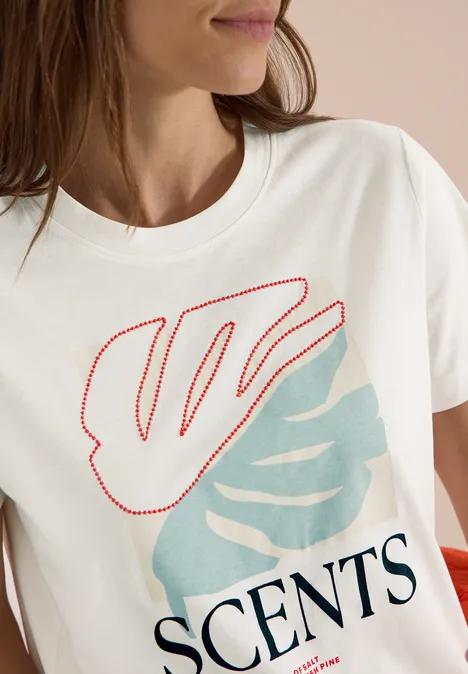 Een vrouw draagt een wit T-shirt met rode en lichtblauwe bladmotieven en de tekst 'SCENTS'.