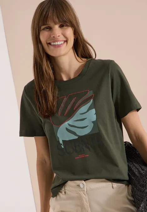 Persoon draagt een kaki t-shirt met bladprint en een beige broek, houdt een donkere trui vast.