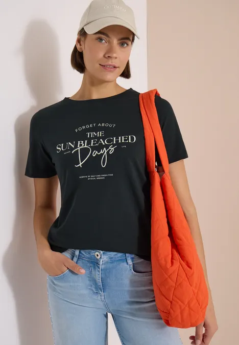 Personne en t-shirt noir, jean bleu clair, sac matelassé orange et casquette devant un mur blanc.