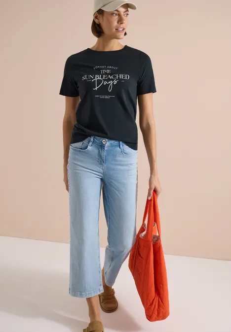 Femme en t-shirt noir, jean bleu clair et chaussures marron, portant un sac orange devant un mur beige.