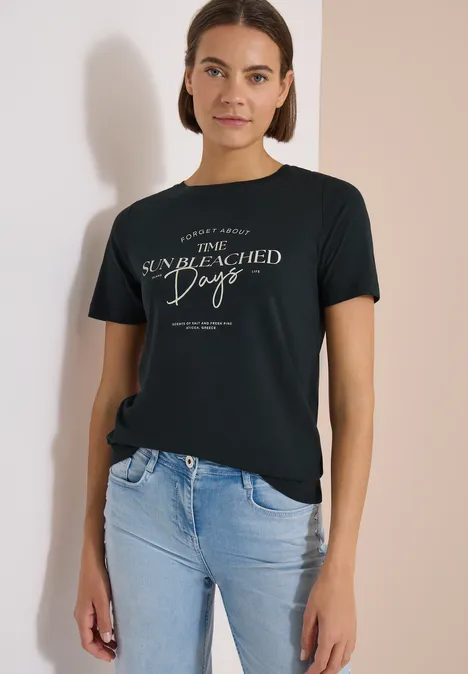 Une femme en jean et t-shirt noir avec un motif blanc, devant un fond clair.