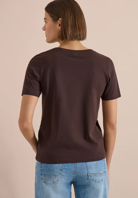 Eine von hinten gesehene Person trägt ein braunes T-Shirt und blaue Jeans vor beigem Hintergrund.