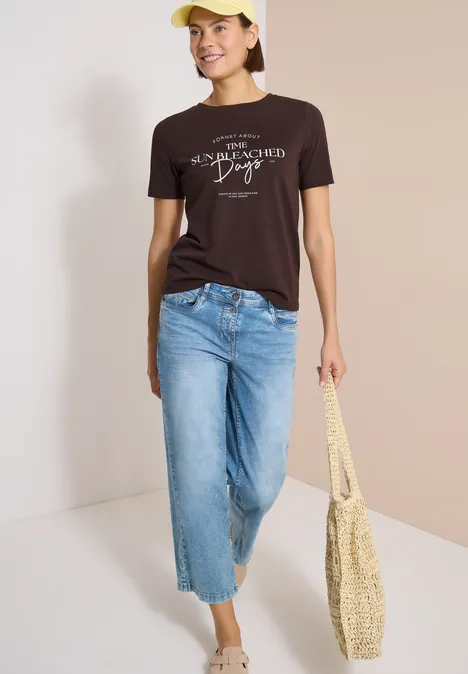 Person mit braunem T-Shirt, hellen Jeans, gelber Mütze und Strohtasche vor hellem Hintergrund
