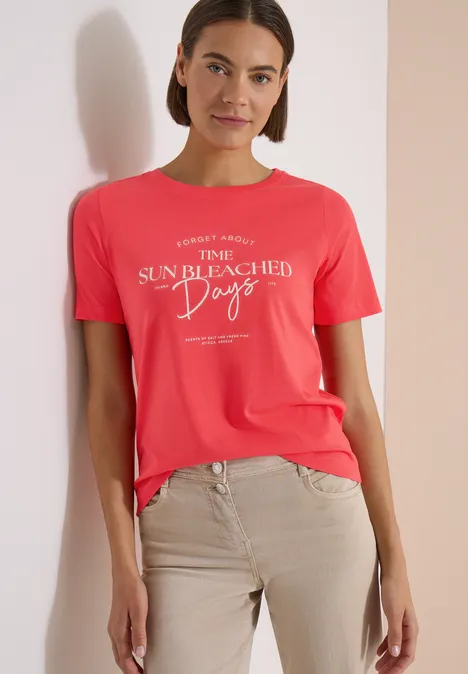 T-Shirt mit Boat Neck und Wording Artwork coral Frau im roten T-Shirt mit Aufschrift und beigen Hosen vor hellem Hintergrund.