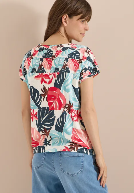Frau von hinten in kurzärmeligem Shirt mit buntem Blattmuster (rot, türkis) und hellblauen Jeans.