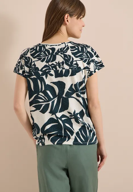 Rückansicht einer Frau in weißem T-Shirt mit dunklem Blattmuster und grüner Hose.